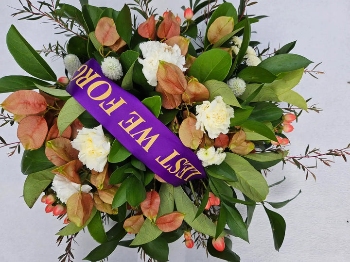 White carnations, dried paper daisies, and green foliage in a sympathy wreath with purple &