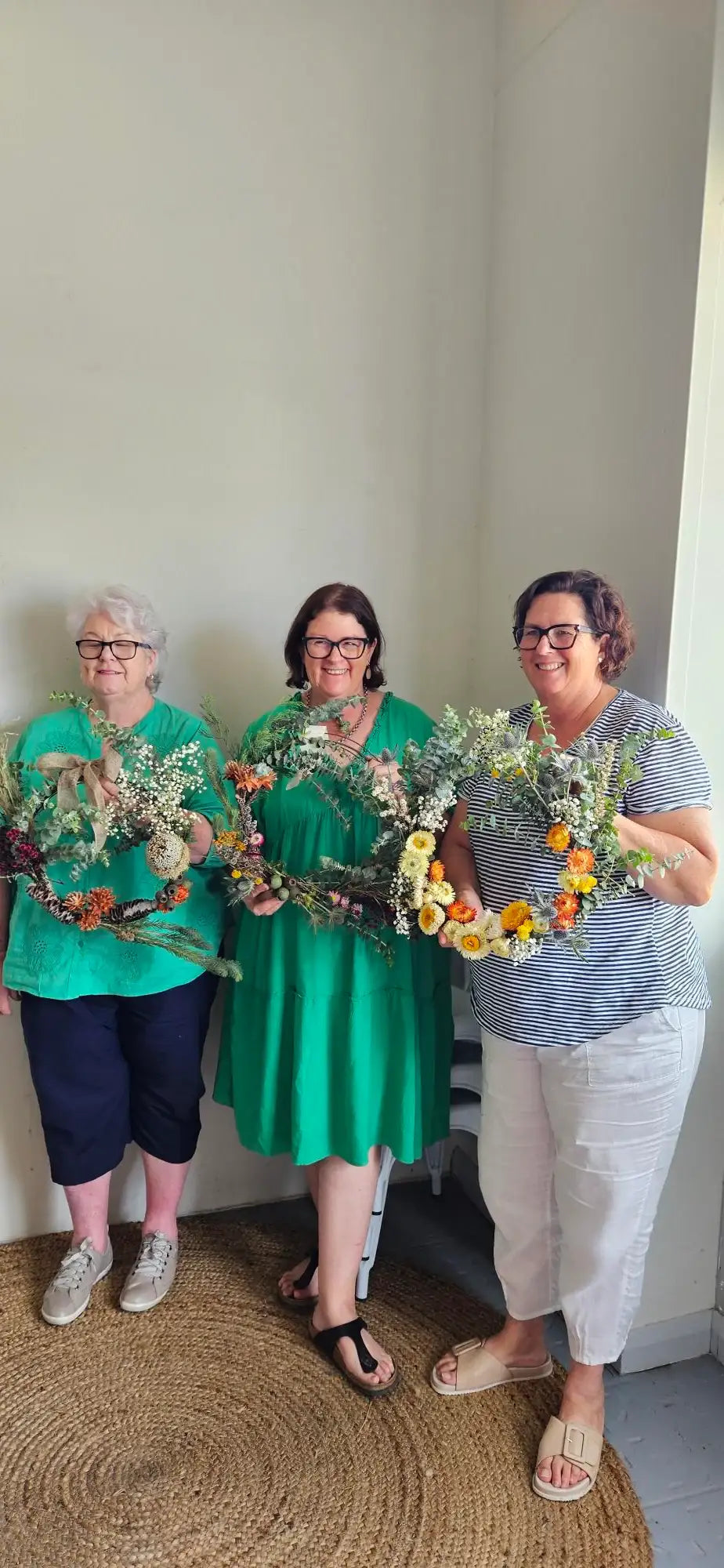 Three women holding handmade floral wreaths with paper daisies and eucalyptus, perfect for garden decor