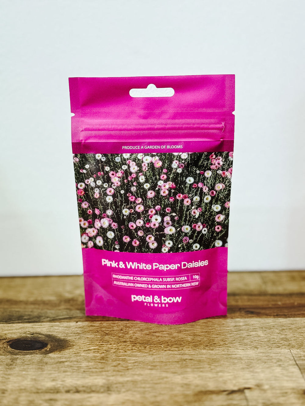 Packaging of Petal and Bow flowers pink and white everlasting paper daisiy seed packet on a wooden surface with a white background